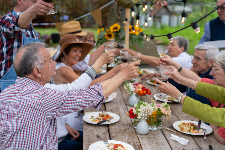 Group of senior friends raising glasses in a toast during an outdoor dinner party. Elderly people enjoying meal and wine in garden with flowers and string lights, celebrating life and friendship.